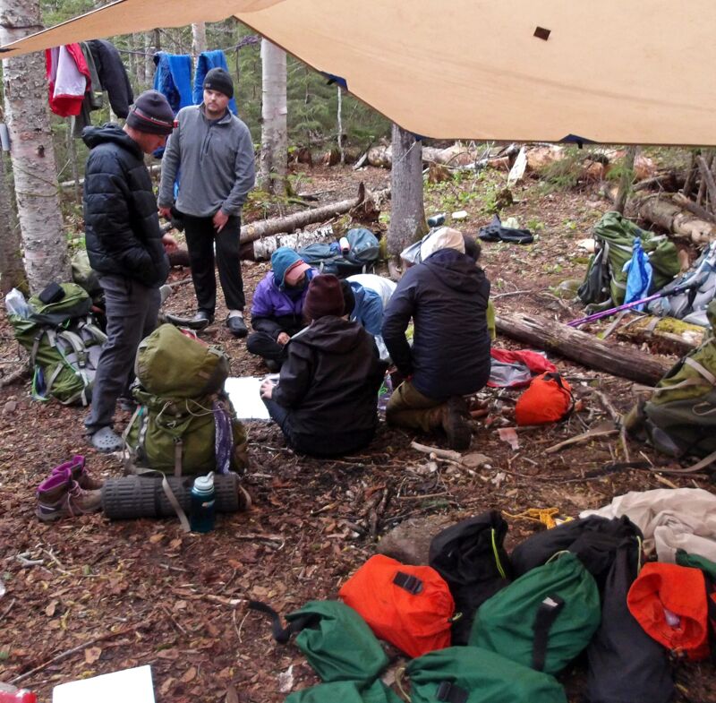 A group of people are gathered under a large tarp in a wooded area, possibly camping. Backpacks and gear are scattered around them on the ground. One person is standing while others are sitting or kneeling, seemingly looking at a map or document. The scene suggests an outdoor adventure or expedition.